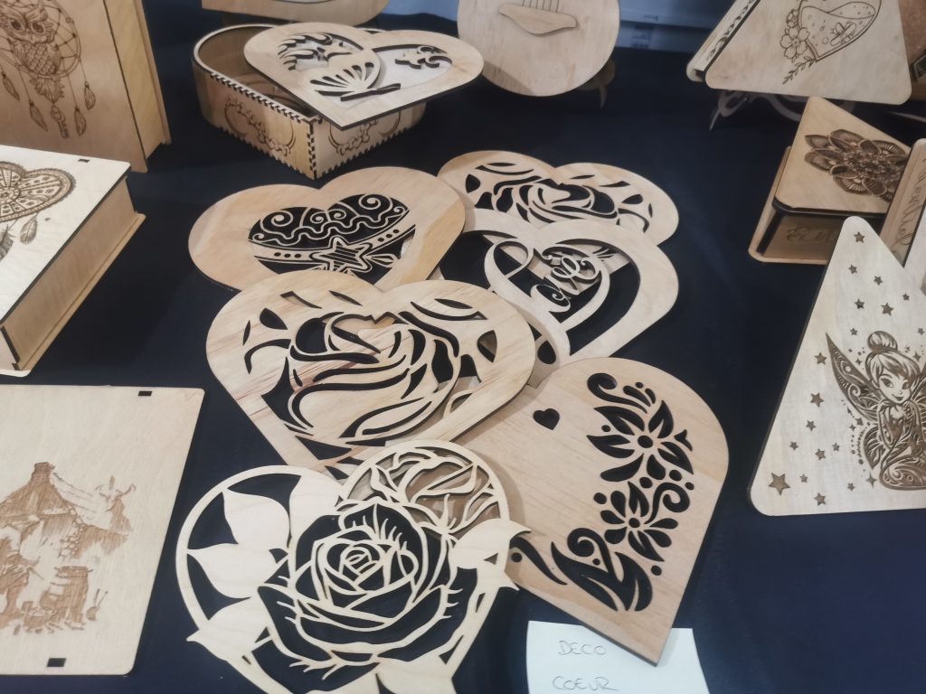 Décorations artisanales en forme de cœur, finement découpées et gravées au laser dans du bois clair, exposées sur un stand.